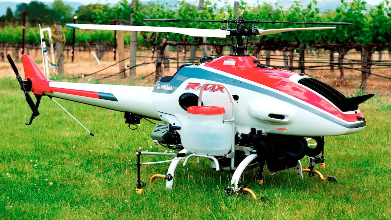 A red and white agricultural drone, the Yamaha RMAX, hovering over a green field with vineyards in the background.